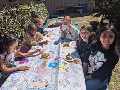 Six children smiling around a long outdoor table covered in a colouring-page tablecloth at a SiW spring camp