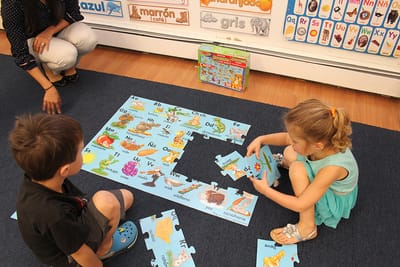 Two children assembling a giant Spanish alphabet floor puzzle with their teacher nearby