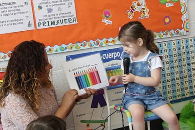 A preschooler holding a microphone and naming her favourite colour in Spanish while her teacher holds up a colour chart