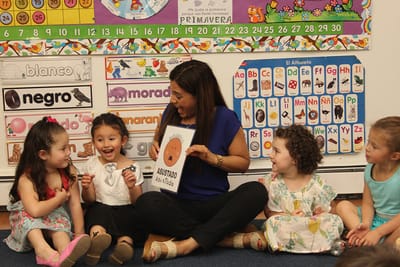 A teacher showing an emotions flashcard to a circle of children in a classroom lined with Spanish colour and alphabet charts